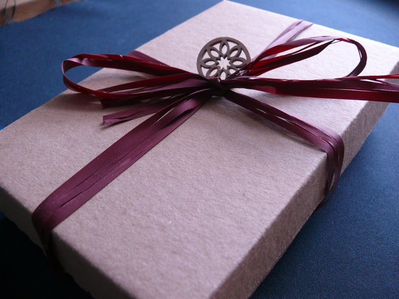 May include: A beige rectangular gift box tied with a deep burgundy ribbon. A decorative wooden embellishment is centered on the ribbon. The box is on a dark blue surface, suggesting a presentation or display.