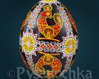 Peut inclure: Un œuf décoratif peint à la main, avec un fond marron foncé. L'œuf présente des motifs complexes en rouge, jaune et blanc, notamment des oiseaux stylisés, des éclats de soleil et des motifs géométriques. L'œuf est présenté sur un support noir.