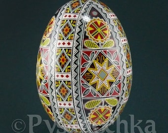May include: A hand-painted Easter egg with intricate geometric patterns in red, yellow, black, white, and blue. The egg is on a stand and has a glossy finish.