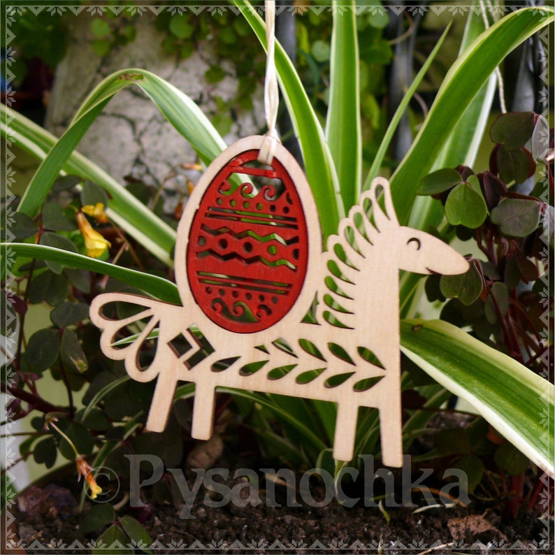 May include: Wooden Easter ornament featuring a horse shape with a red egg design. The egg has decorative cutouts. The ornament is made of light-colored wood and is hanging from a string. The word "Pysanochka" is visible.