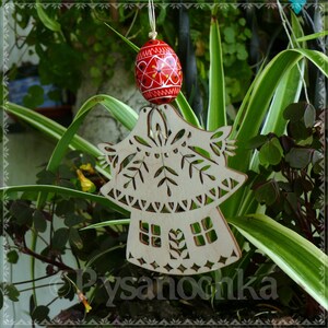 May include: A wooden ornament with a cut-out design of a house and birds, paired with a red and yellow painted Easter egg. The egg is suspended above the wooden ornament. The background features green plants.