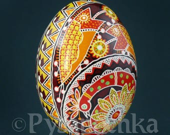 Puede incluir: Un huevo de Pascua pintado a mano con diseños intrincados en tonos de amarillo, naranja, rojo y marrón. El huevo presenta patrones geométricos, motivos florales y un pájaro estilizado.