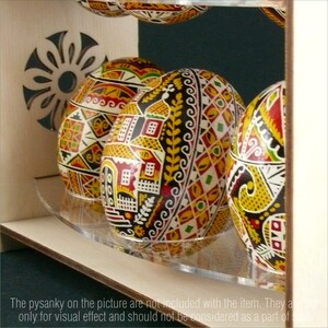 Puede incluir: Un expositor de madera con un estante de acr&iacute;lico transparente para exhibir huevos decorativos. Los huevos est&aacute;n pintados con intrincados patrones en rojo, amarillo, negro y blanco.