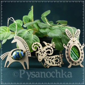 May include: Three wooden Easter ornaments, a horse, a rooster, and a bunny, each holding a decorated egg. The ornaments are cut out and have intricate designs. The background is a green plant.