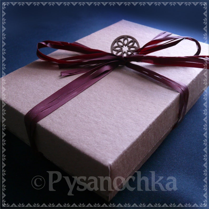 May include: A brown gift box with a burgundy ribbon and a wooden flower accent. The box is sitting on a blue surface.