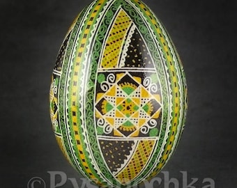 Puede incluir: Un huevo de Pascua pintado a mano con intrincados patrones geométricos en negro, verde, amarillo y blanco. El huevo está decorado con un motivo central de un cuadrado que contiene una estrella.