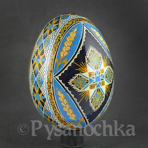 May include: A hand-painted pysanka egg with intricate geometric and floral designs. The egg features a dark blue background with patterns in yellow, white, and light blue. The egg is standing on a black stand.
