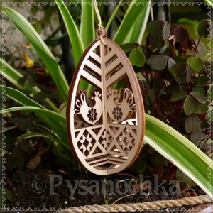 May include: Wooden Easter egg ornament with a laser-cut design. The ornament features two stylized roosters, floral details, and geometric patterns. The egg is suspended by a natural twine string, perfect for Easter decor.