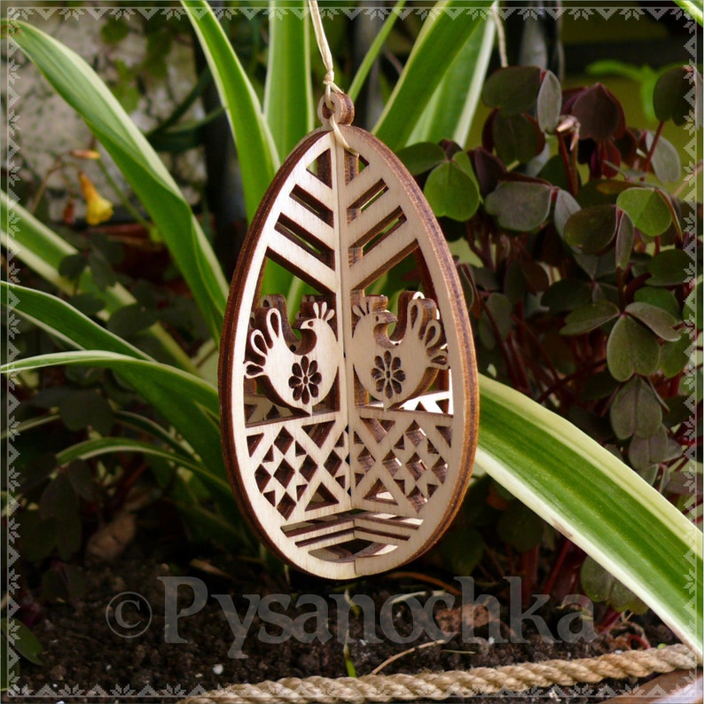 May include: Wooden Easter egg ornament with a laser-cut design. The ornament features two stylized roosters, floral details, and geometric patterns. The egg is suspended by a natural twine string, perfect for Easter decor.