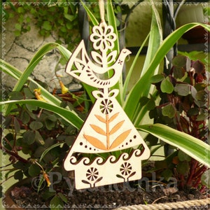 May include: Wooden ornament featuring a bird and floral design. The ornament is light brown with cut-out details, including a flower, bird, and stylized leaves. The ornament is hanging in front of green plants.