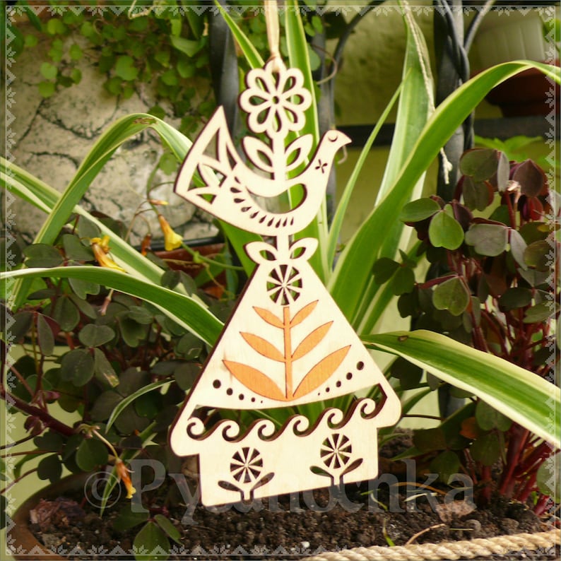 May include: Wooden ornament featuring a bird and floral design. The ornament is light brown with cut-out details, including a flower, bird, and stylized leaves. The ornament is hanging in front of green plants.