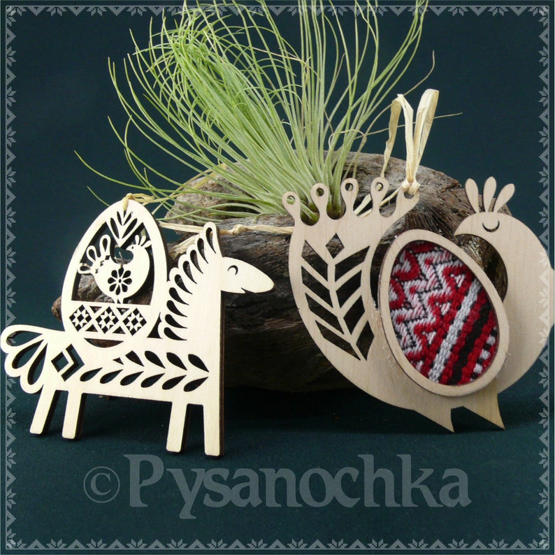 May include: Three wooden ornaments in the shape of a horse, a bird, and an egg. The horse and bird have intricate cutouts and the egg has a red and white embroidered design. The ornaments are all hanging from a string.