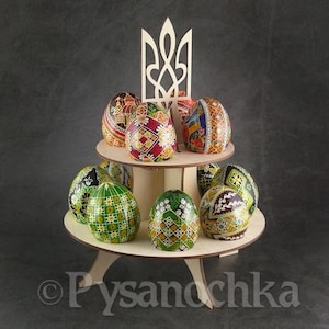 May include: A wooden stand with a Ukrainian trident design on top, holding a collection of intricately painted Easter eggs. The eggs are decorated with various patterns and colors, including red, green, yellow, and blue.