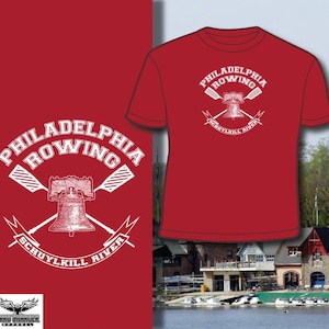 May include: Red t-shirt with a white graphic featuring the words "Philadelphia Rowing" above a bell and crossed oars, with "Schuylkill River" below. The design is repeated on the left side of the image. The shirt is displayed against a background of a river and buildings.