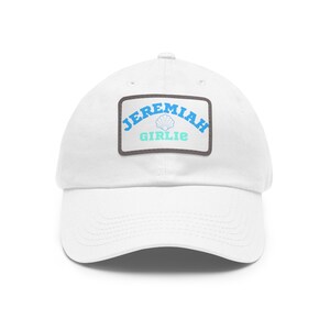 May include: A white baseball cap featuring a rectangular patch on the front. The patch has a dark gray border and displays the text JEREMIAH in blue, a light blue seashell icon, and the word GIRLIE in light green lettering on a white background.