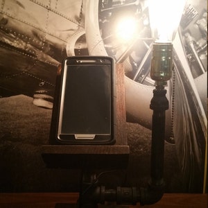 May include: A rustic wooden phone stand with a black phone resting on it. The stand is attached to a black metal pipe lamp with a light bulb.