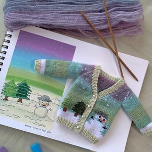 Może przedstawiać: Miniaturowy, ręcznie robiony sweter w kolorach zieleni, fioletu i bieli, ozdobiony wzorem bałwana i drzew. Obraz zawiera szkic zimowej scenerii, włóczkę, druty do robienia na drutach i markery, sugerując projekt rzemieślniczy.