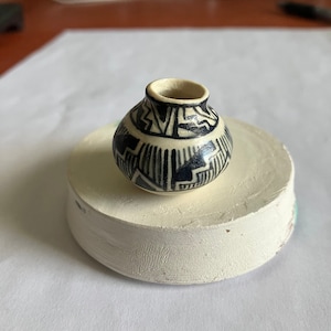 Peut inclure: Petit pot en céramique crème, de forme arrondie et à ouverture étroite. Il est orné d'un motif géométrique bleu foncé composé de lignes et de formes. Le pot repose sur une base circulaire blanche. Le design est symétrique.