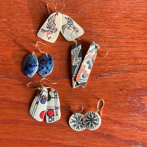 May include: A collection of handmade earrings with various designs. Styles include bird motifs, abstract patterns, and geometric shapes. The earrings feature gold-colored hooks and are displayed on a wooden surface.