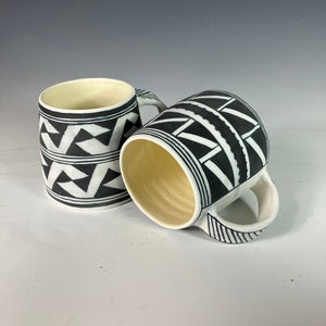 Peut inclure: Deux mugs en céramique avec des motifs géométriques noirs et blancs. Les mugs ont une forme cylindrique avec une anse et un intérieur crème. L'extérieur présente un motif répétitif de triangles et de lignes.