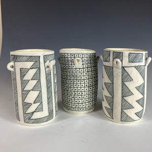Pode incluir: Três vasos cilíndricos de cerâmica com padrões geométricos em preto e branco. Os vasos têm pequenas alças de laço e designs variados, incluindo triângulos, losangos e um padrão de grade. Os vasos são exibidos em um fundo neutro.