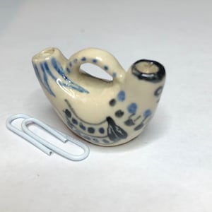 Puede incluir: Una pequeña jarra de cerámica blanquecina con asa y dos picos. Decorada con diseños florales azules y negros. Un clip blanco está al lado de la jarra.