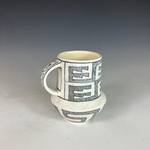 Peut inclure: Une tasse en céramique blanche avec un motif géométrique noir. La tasse a une anse et une base arrondie. L'intérieur de la tasse est de couleur crème. Le motif est répété sur la anse et la base.