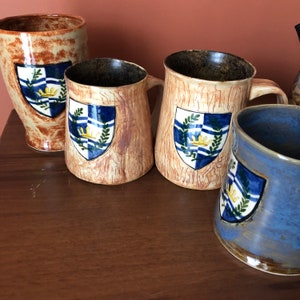 Puede incluir: Colección de tazas y un jarrón de cerámica hechos a mano. Las tazas son beige con textura de vetas de madera y un diseño de escudo azul y blanco. Una taza es azul. El jarrón es beige con vetas marrones y un escudo. Las tazas tienen asas.