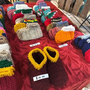 May include: A variety of colorful crocheted slipper socks for sale. The socks are arranged in rows on a red tablecloth. The price tag reads $10.00.  The socks are labeled "Sm Kids" and "Toddler".