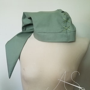 May include: A sage green fabric hood with a long, flowing tail. The hood has a seam down the back and is secured with a series of X-shaped stitches.