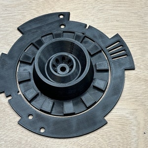 May include: A black 3D printed plastic part with a central circular opening and a series of concentric rings around it.