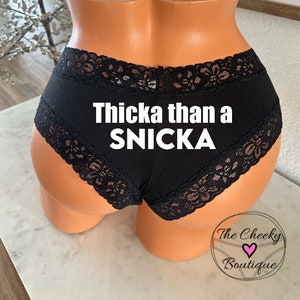 May include: Black underwear with lace detailing, displayed on a mannequin. The text "Thicka than a SNICKA" is printed on the front. The Cheeky Boutique logo is visible at the bottom.