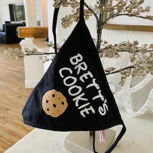 May include: Black fabric face mask with white text that says "Brett's Cookie" and a brown cookie graphic.
