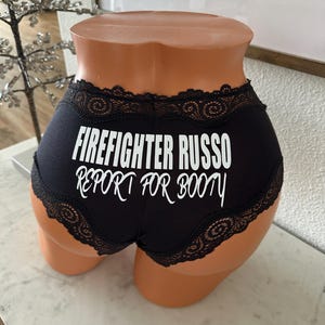 May include: Black underwear with a lace trim and the text "FIREFIGHTER RUSSO REPORT FOR BOOTY" in white. The underwear is displayed on a mannequin.