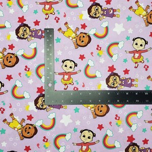 Peut inclure: Un tissu violet clair avec un motif r&eacute;p&eacute;t&eacute; de personnages de dessins anim&eacute;s, d'arcs-en-ciel, d'&eacute;toiles et de notes de musique. Les personnages sont dans diff&eacute;rentes poses, portant diverses tenues. Une r&egrave;gle est plac&eacute;e sur le tissu.