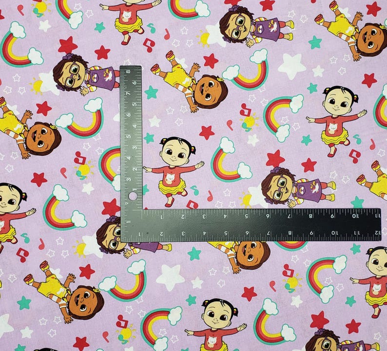 Peut inclure: Un tissu violet clair avec un motif r&eacute;p&eacute;t&eacute; de personnages de dessins anim&eacute;s, d'arcs-en-ciel, d'&eacute;toiles et de notes de musique. Les personnages sont dans diff&eacute;rentes poses, portant diverses tenues. Une r&egrave;gle est plac&eacute;e sur le tissu.