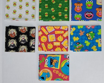 May include: Seven folded fabric squares featuring Sesame Street characters. Patterns include Kermit the Frog on white and green, various characters on blue, Statler and Waldorf on black, Big Bird on red, puzzle pieces on blue, and Cookie Monster on pink.