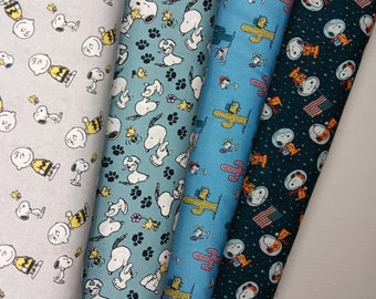 May include: Four rolls of fabric with cartoon character prints. The fabrics feature Snoopy, Charlie Brown, and Woodstock in various designs. Colors include light gray, teal, light blue, and dark teal. Patterns include paw prints, cacti, and Snoopy in an astronaut suit.