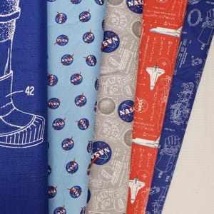 May include: A collection of NASA-themed fabric rolls in various colors. Designs include the NASA logo, space shuttles, and technical diagrams. Colors include blue, light blue, gray, and red. The number "42" is visible on one of the rolls.