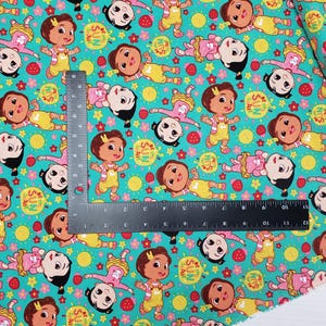 Peut inclure: Tissu turquoise avec un motif r&eacute;p&eacute;t&eacute; de personnages de dessins anim&eacute;s en salopettes jaunes et robes roses. Le motif comprend des soleils, des fraises et des fleurs. Le texte "SHINE" et "SUN" est &eacute;galement visible.