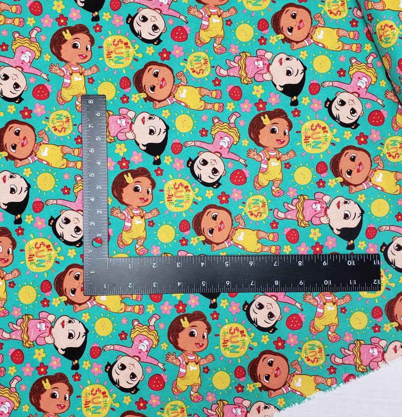 Peut inclure: Tissu turquoise avec un motif r&eacute;p&eacute;t&eacute; de personnages de dessins anim&eacute;s en salopettes jaunes et robes roses. Le motif comprend des soleils, des fraises et des fleurs. Le texte "SHINE" et "SUN" est &eacute;galement visible.