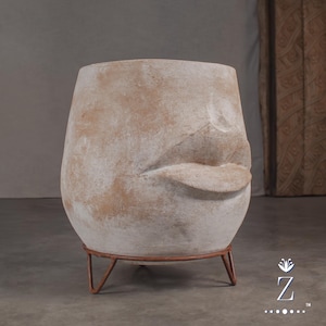 May include: A large, rounded planter with a weathered, off-white and tan textured finish. The exterior features a sculpted relief of a nose and full lips. The vessel is supported by a thin, circular, rust-coloured metal stand with three legs.