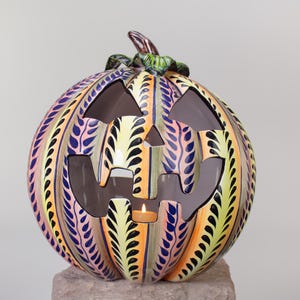 Peut inclure: Une citrouille en céramique en forme de jack-o'-lantern avec un visage sculpté et une bougie à l'intérieur. La citrouille est décorée de rayures colorées et de motifs de feuilles dans des tons de jaune, orange, rose, bleu et noir.