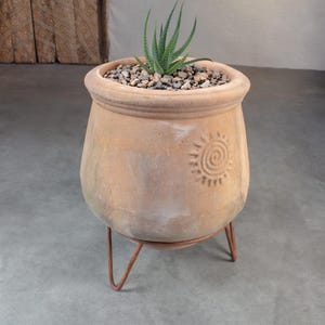 May include: A light brown terracotta planter with a rounded body and a thick rim, featuring an embossed sun design on the side. The planter sits on a thin, three-legged copper-colored metal stand and contains a small green succulent in gravel.
