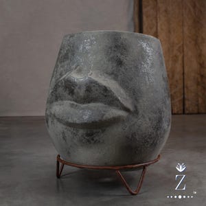 May include: A grey, textured, rounded planter featuring a sculpted human nose and lips on the front surface. The vessel is supported by a thin, three-legged, rust-coloured metal stand that elevates the base slightly above the ground surface.