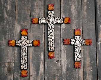 May include: Three decorative wooden crosses with intricate designs and a flame-like motif at the top and bottom. The crosses are black with silver and white embellishments and feature orange and red accents. They are displayed against a weathered gray wooden background.