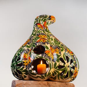May include: A ceramic gourd-shaped candle holder with a green and white background and orange and black butterfly designs. The candle holder has cut-out designs and a lit candle inside.