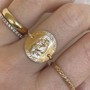 May include: A collection of gold rings, including a wide band, a diamond-studded band, and a delicate ring with a curved design. A gold pendant ring features an engraved image and the text "SAINT ANTHONY FOR US".
