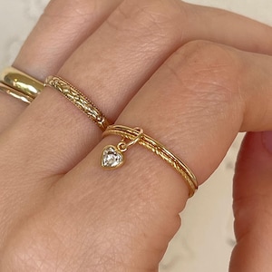 May include: A hand wearing several gold rings. One ring has a heart-shaped charm with a clear stone. Other rings include a plain band and a textured band. The rings are displayed against a light background.