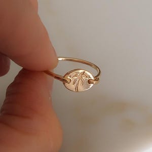 May include: A gold-toned ring featuring a thin, circular band connected to a small, horizontal oval-shaped charm. The center of the oval charm is engraved with a detailed palm tree design, showcasing a smooth, polished metallic finish throughout.